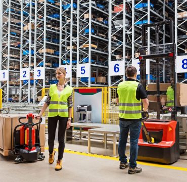 Two Storehouse employees in uniform standing near pallet truck and forklift in modern automatic warehouse.Boxes are on the shelves of the warehouse. Warehousing, machinery concept. Logistics in stock.